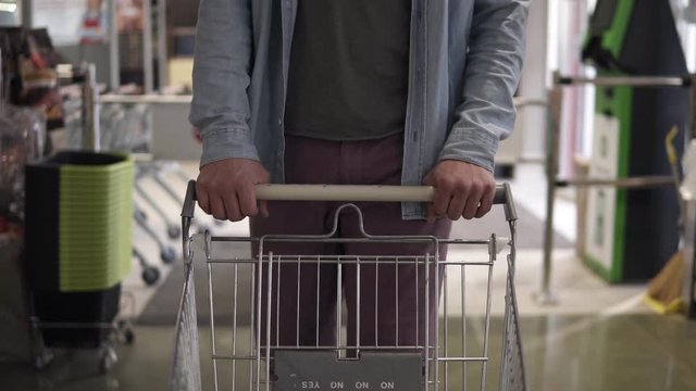Cropped front footage of a man in blue shirt and violet pants pushing the shopping empty shopping trolley the supermarket entry. The guy came for shopping to a big mall