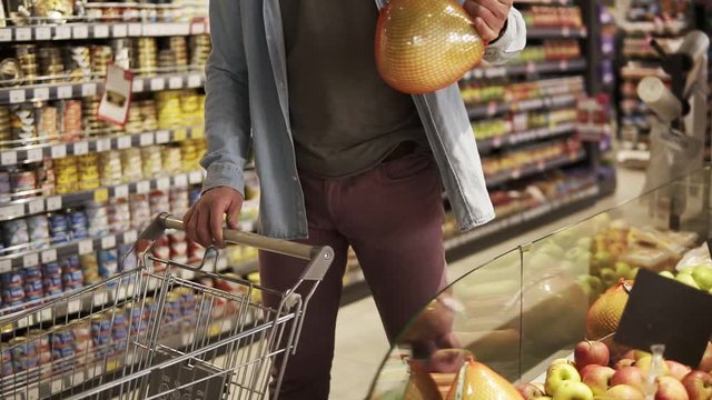 Cropped footage of unrecognizable man in blue shirt shopping for fruits and vegetables in produce department of a grocery store supermarket. Taking pomelo fruit, putting it into shopping trolley and