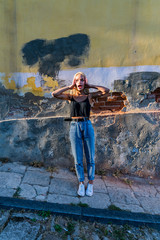 Excited blonde teen girl in black top and blue jeans stands on the sidewalk and holds her hands on her head, old cracked wallon the background