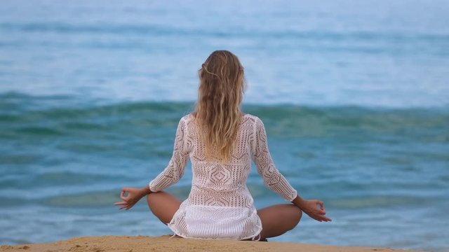 yoga woman practices yoga and meditates in lotus position on beach sea. healthy female girl sits in pose of lotus sukhasna meditating or practicing yoga in nature ocean outdoors, relaxing 4 K slow-mo