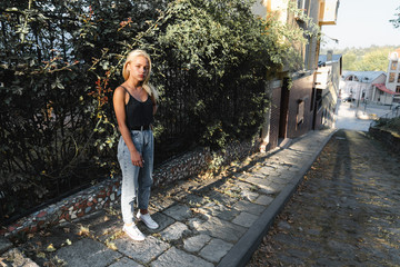 Fototapeta na wymiar Attarctive calmy blonde girl in casual clothes stands near the green fence among the old city street