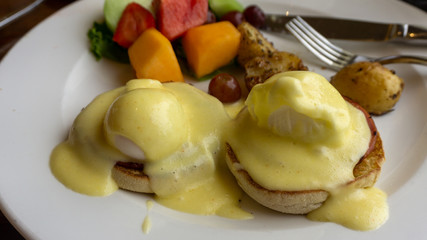 Close up of eggs benedict with two eggs and bacon on muffins with hollandaise sauce and pineapples