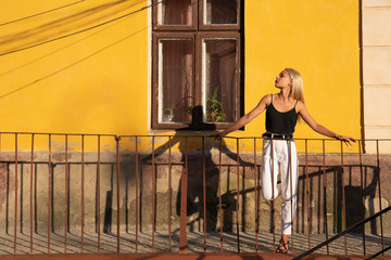 Fototapeta na wymiar Trendy blonde girl in casual clothes leaned on the old balcony railing, yellowe wall with old wooden window on the background