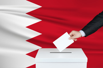 Election in Bahrain. The hand of man putting his vote in the ballot box. Waved Bahrain flag on background.