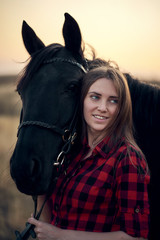 Fototapeta premium lovely young girl in a plaid shirt hugs a black horse at sunset in the mountains