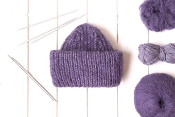 Purple hat and yarn balls, metal needles on the white wooden background. 