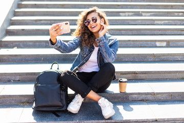 Portrait of young beautiful woman sitting on stairs and taking selfie photo with phone