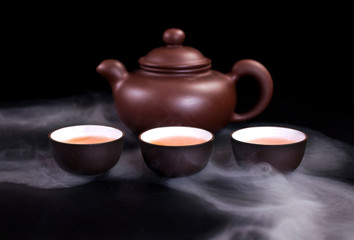 Asian tea ceremony. Chinese clay teapot and ceramic tea cups with vapor on a black background.