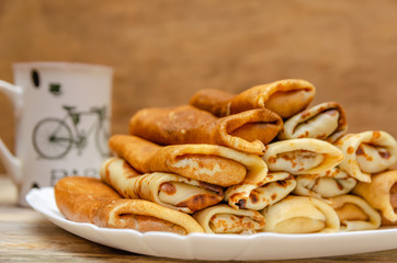 Pancakes with filling are a pyramid on a white plate standing on a wooden background close-up, in the background a Cup