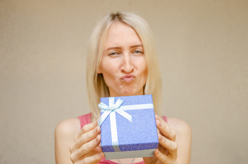 blonde girl pranks, grimaces and holds a blue shiny gift box in front of her, copy space. Focus on the present. concept of the celebration.