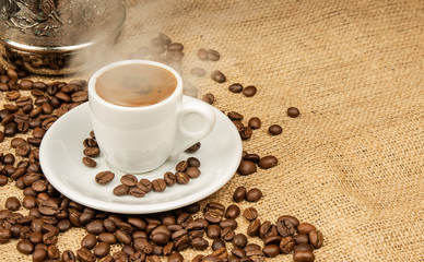 Cup of Greek coffee on sack background with smoke. Turkish coffee in white small mug and coffee beans. Traditional Greek coffee concept. Copy space for text.