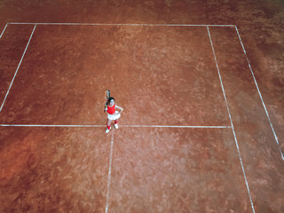 A young woman with a tennis racket in her hand stands in the center of a tennis court and looks into the camera. Top view, aerial view