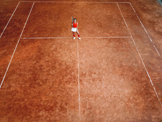 A young woman with a tennis racket in her hand stands in the center of a tennis court and looks into the camera. Top view, aerial view