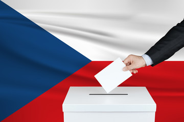 Election in Czech Republic. The hand of man putting his vote in the ballot box. Waved Czech Republic flag on background.