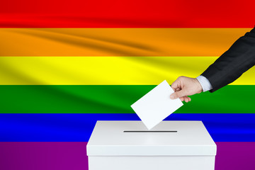 Election in Lgbt. The hand of man putting his vote in the ballot box. Waved Lgbt flag on background.