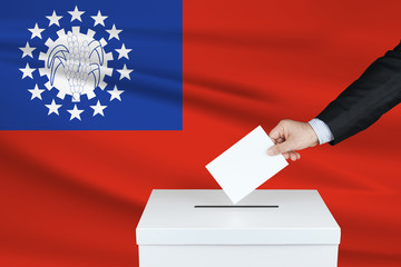 Election in Myanmar. The hand of man putting his vote in the ballot box. Waved Myanmar flag on background.