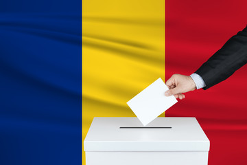 Election in Romania. The hand of man putting his vote in the ballot box. Waved Romania flag on background.