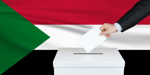 Election in Sudan. The hand of man putting his vote in the ballot box. Waved Sudan flag on...
