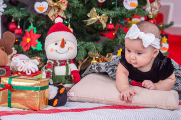 A white baby in Christmas tree decoration with black dress and white bow, leaning on her hands looks at something