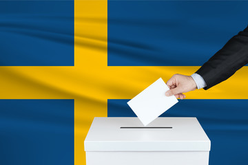 Fototapeta premium Election in Sweden. The hand of man putting his vote in the ballot box. Waved Sweden flag on background.