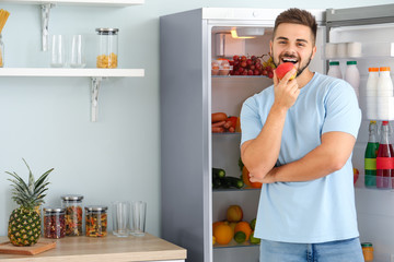 Young man with apple near open fridge in kitchen