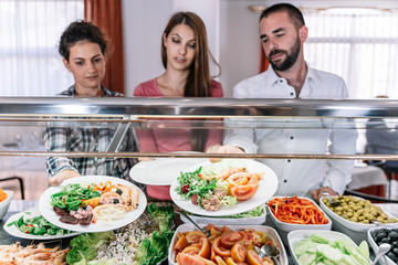 People with dishes in their hands waiting to be served at a self-service restaurant counter.