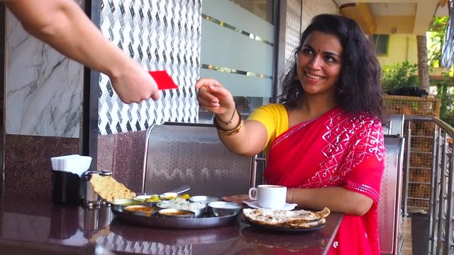 beautiful woman pay with credit card for dosa chutney naan and indian tali in a restaurant.She wearing red sari with gold earrings , with tilaka on her forehead.