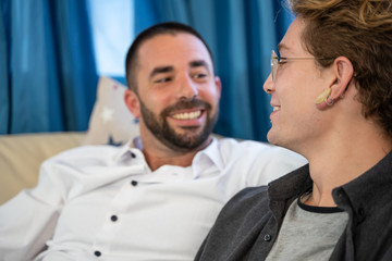 Young guy with piercings and glasses looking at his gay partner on a sofa