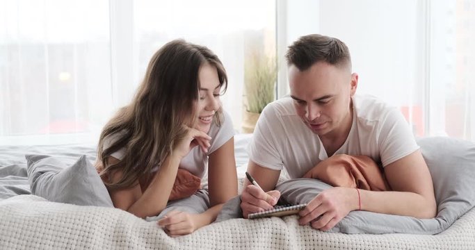 Young couple writing in notepad lying on bed at home