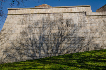 Fragment of an old fortified wall of stone blocks. There is a shadow from a tree without leaves and green grass at the foot. Citadel in the city of Besancon. France. Background.