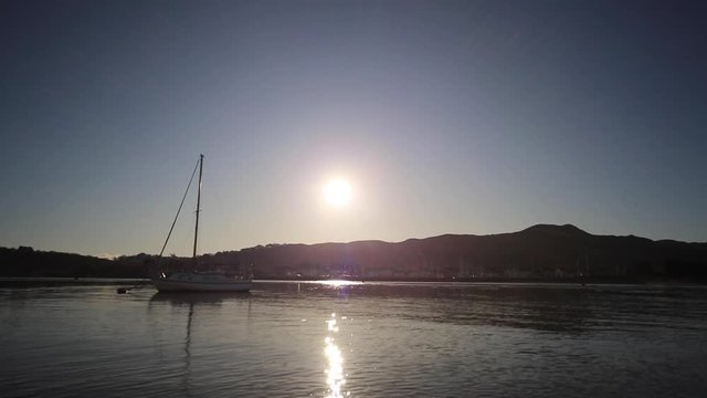 Wide angle view of a yacht bobbing up and down gently on its mooring buoy as the sun sets behind the mountains in the background
