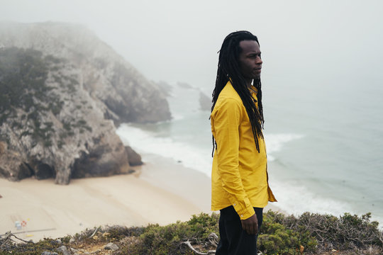 African male wearing long dreadlocks standing on ocean shore, white waves and cliffs in fog on background