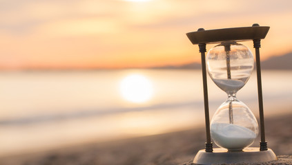 Hourglass in the dawn time. Sand passing through the glass bulbs of an hourglass measuring the passing time as it counts down to a deadline or closure on a sunset/ sunrise beach background.  - obrazy, fototapety, plakaty