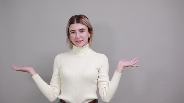 Caucasian attractive girl over gray wall making unimportant gesture while lifting the shoulders wearing white sweater