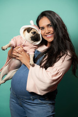 Happy pregnant brunette woman with pug in her hands on empty blue background