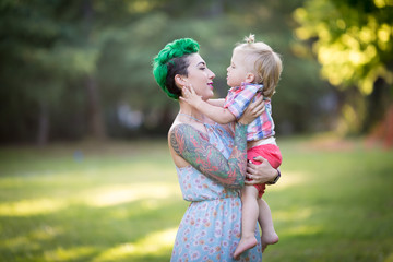 A portrait of Caucasian young woman with green hair and tattoo holding a blond toddler boy in her hands. Hugging, Mother's day concept