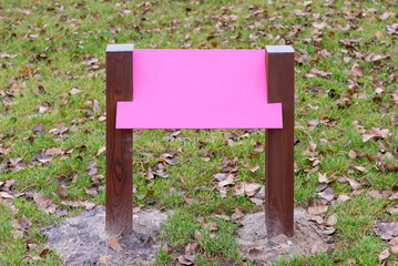 Blank pink wooden board for information and indications in the park, with a background of green grass covered by autumn leaves