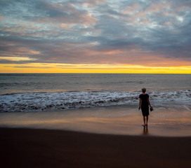 A woman stands on a Kauai beach at sunset, looking at the golden skyline, wet sand at her feet reflecting the soft light.