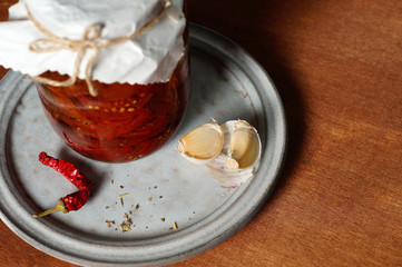 Glass jar with dried tomatoes on a ceramic plate with garlic cloves, Provencal herbs and a pod of hot chili peppers. Copy space, horizontal layout, top view, close-up.