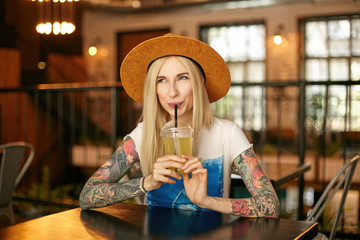 Cheerful blonde tattooed woman with casual hairstyle drinking lemonade while waiting for friends in...