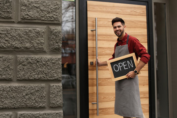 Young male business owner holding OPEN sign near his cafe. Space for text