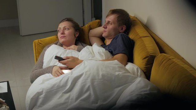 Young couple watching TV talking lay on yellow sofa woman and man covered white blanket middle shot