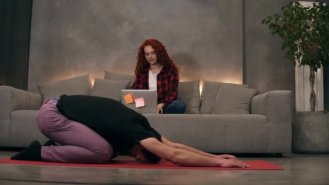 Young caucasian couple at relaxing home living room, curly readheaded firl using modern wireless laptop on a grey couch while her boyfriend is doing yoga, stretching on a red mat. Front view. Loft