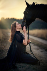 Beautiful young girl in a black dress at sunset kneels next to her black horse and hugs him in the...