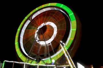 Long Exposure Shot of a well illuminated and colorful Giant Ferris Wheel rotating speedily on its principal axis, during the night hours, in a local fair for the amusement and thrill of visitors.