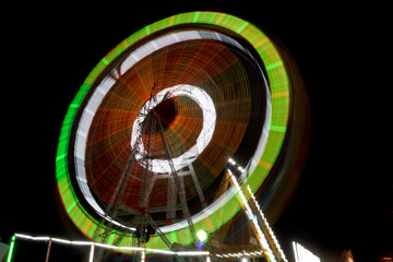 Long Exposure Shot of a well illuminated and colorful Giant Ferris Wheel rotating speedily on its principal axis, during the night hours, in a local fair for the amusement and thrill of visitors.