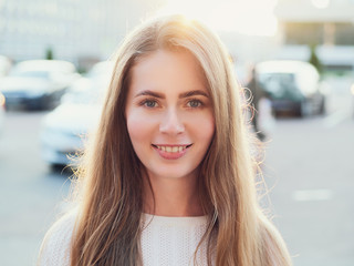 Close up headshot of adorable blonde haired charming smiling woman with blue eyes looking into camera in city street with bright sun flare