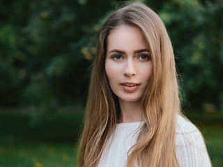 Close up headshot of adorable blonde haired charming smiling woman with blue eyes looking into camera in evening city park