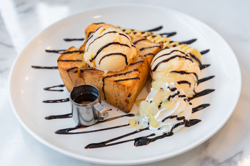 Side view of Honey toast topped vanilla ice cream and chocolate sauce served with banana slices and whipped cream on the white marble table.