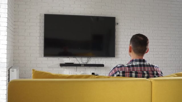 Young modern caucasian man in plaid shirt sitting on the yellow sofa watching plasma TV. Screen is black. Switches channels using the remote. Back view